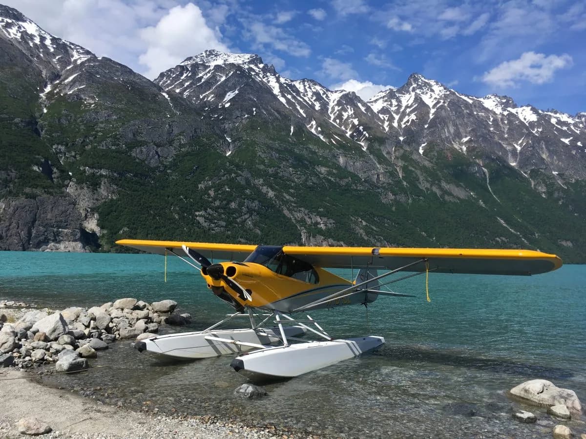 A yellow and grey Carbon Cub floatplane is partially in a turquoise lake, with snow-capped mountains and green trees in the background.