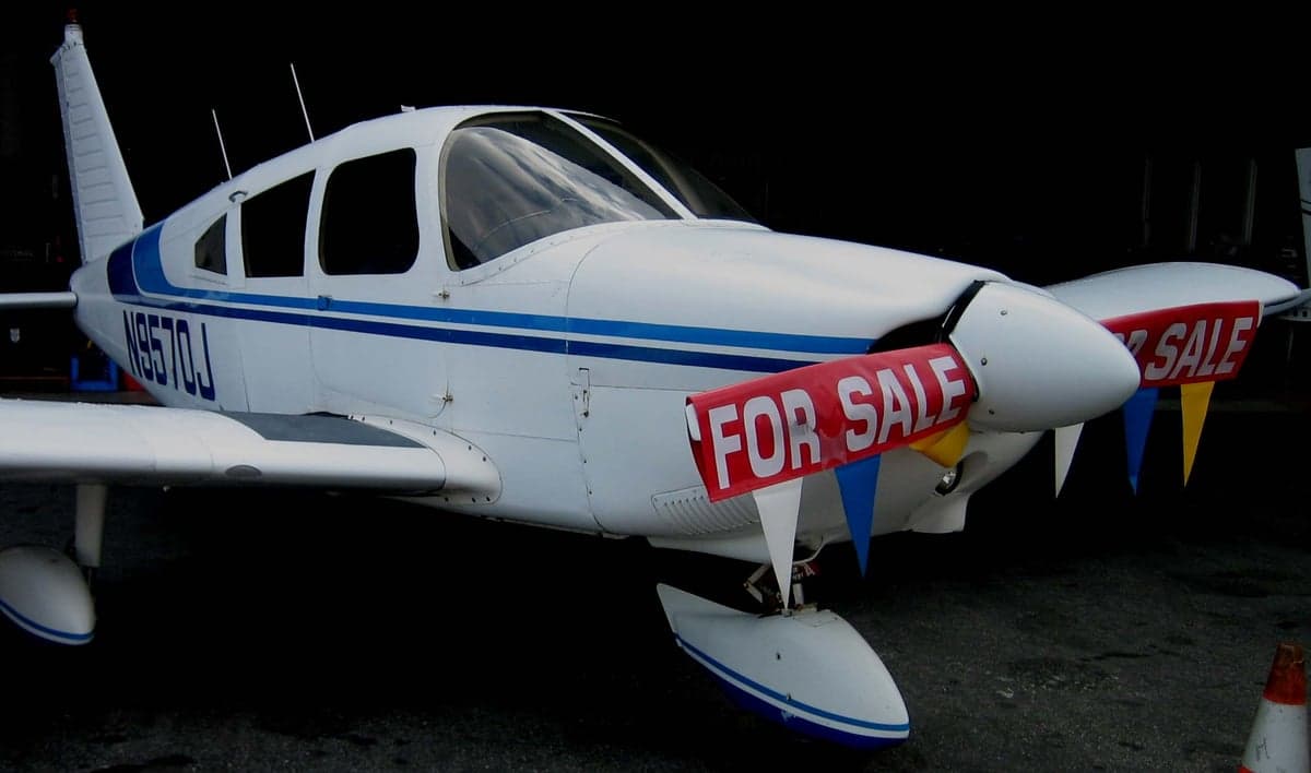 A white and blue Piper aircraft with a 'FOR SALE' banner on the nose.