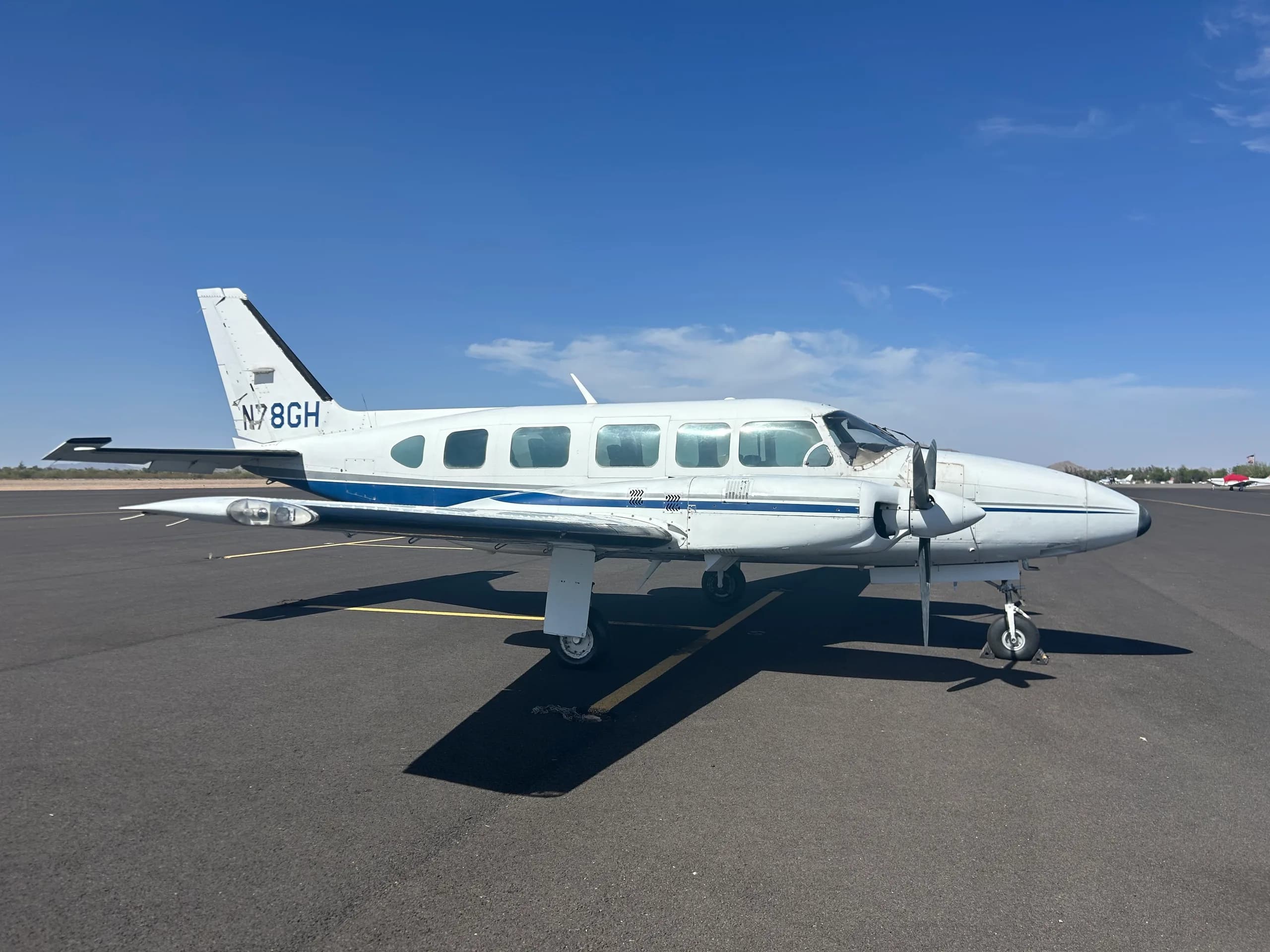 A white twin-engine aircraft with blue and grey stripes, registration N78GH, parked on an asphalt tarmac under a clear blue sky.