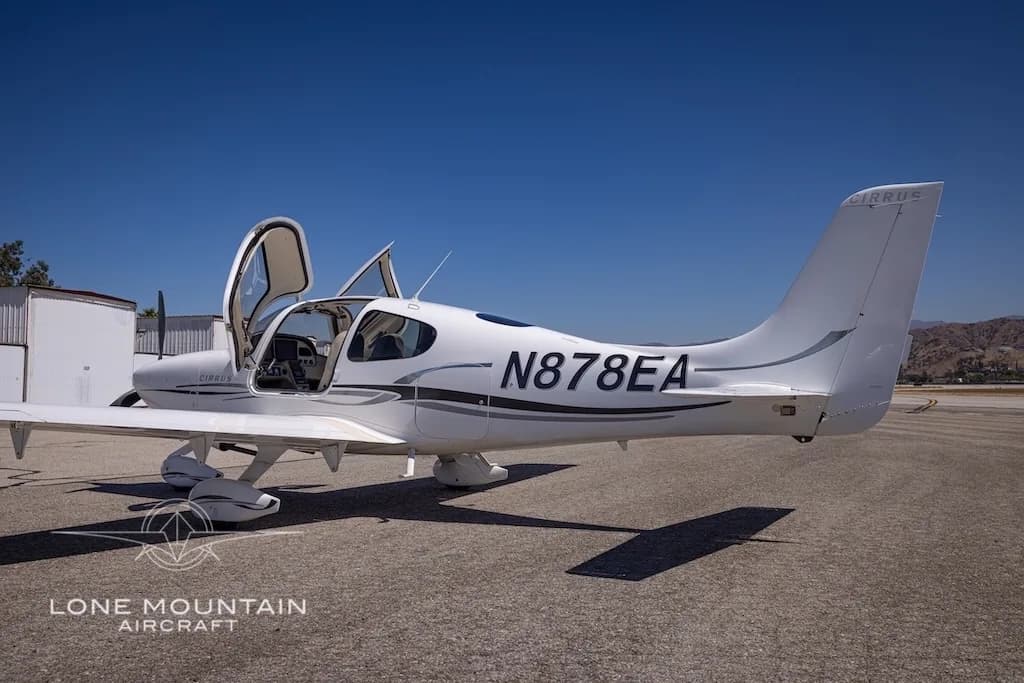 A white Cirrus aircraft with registration N878EA and open doors is parked on an airfield, viewed from the side.