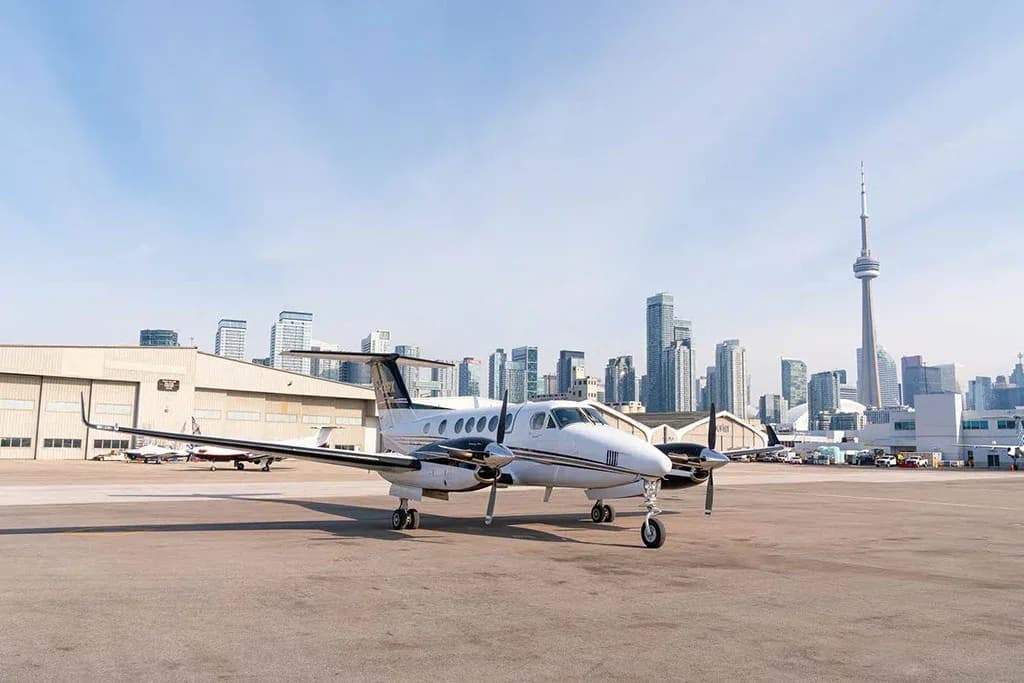 A wide shot of a white and black twin-engine turboprop aircraft (King Air) parked on an airfield with the Toronto skyline in the background.