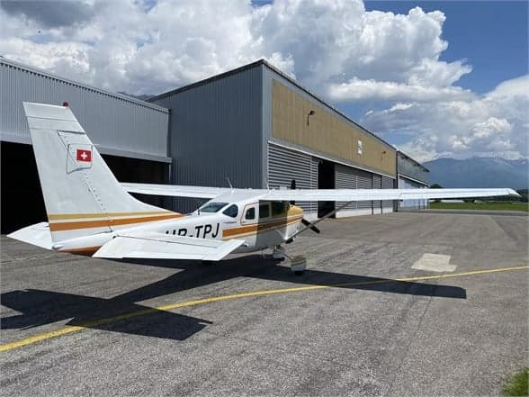 A Cessna 210 Centurion aircraft with a white and gold paint scheme is parked on the tarmac in front of a hangar.