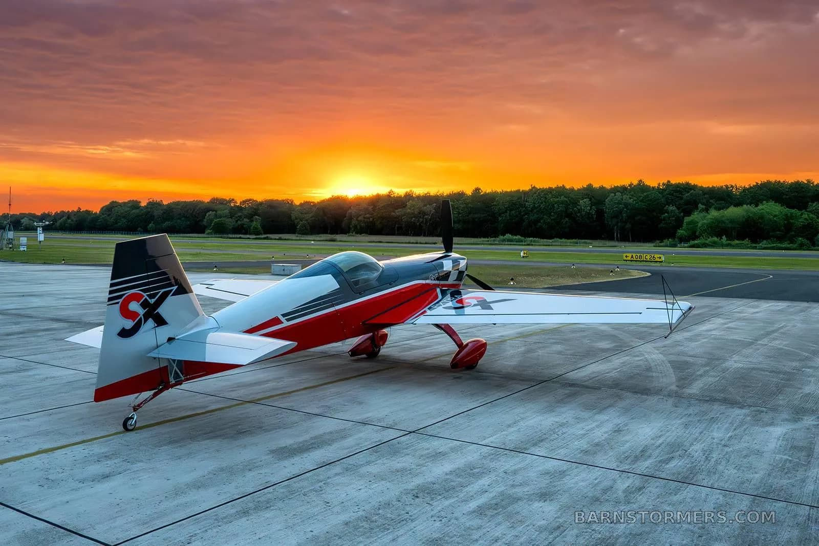 A red, black, and silver Extra 330SC aerobatic aircraft parked on an airport tarmac at sunset with an orange sky.