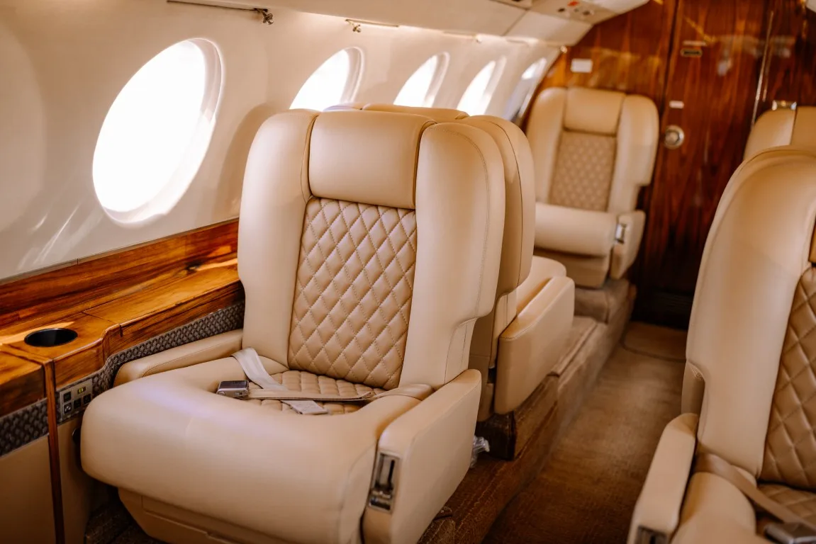 Close-up of tan leather seats with quilted patterns in an aircraft cabin, next to large oval windows.