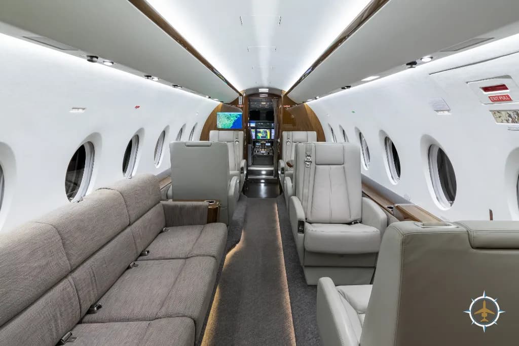 The full length of the business jet cabin, showing a beige sofa on the left and light grey seats on the right, looking towards the cockpit.