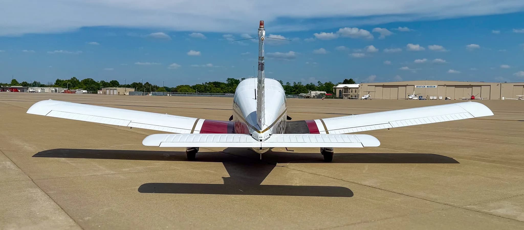 A rear view of the white, red, and gold Piper Cherokee aircraft (N8534N) on an airfield with distant hangars.