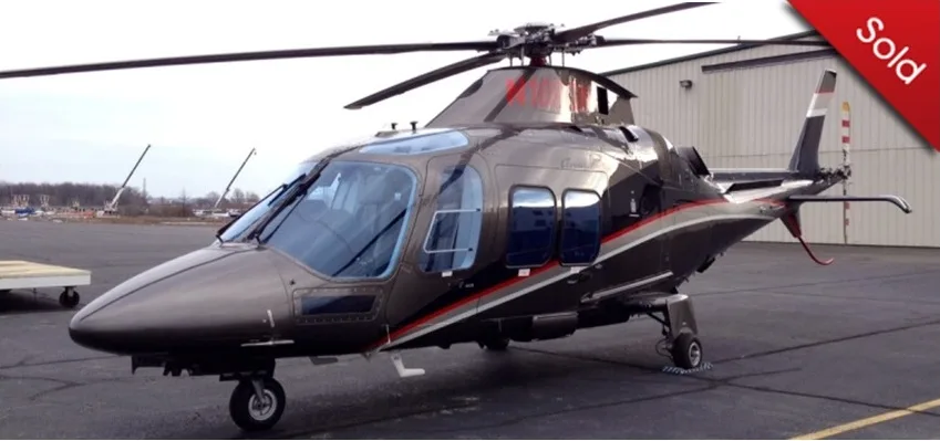 A dark grey executive helicopter with red and silver stripes parked on an asphalt apron in front of a hangar.