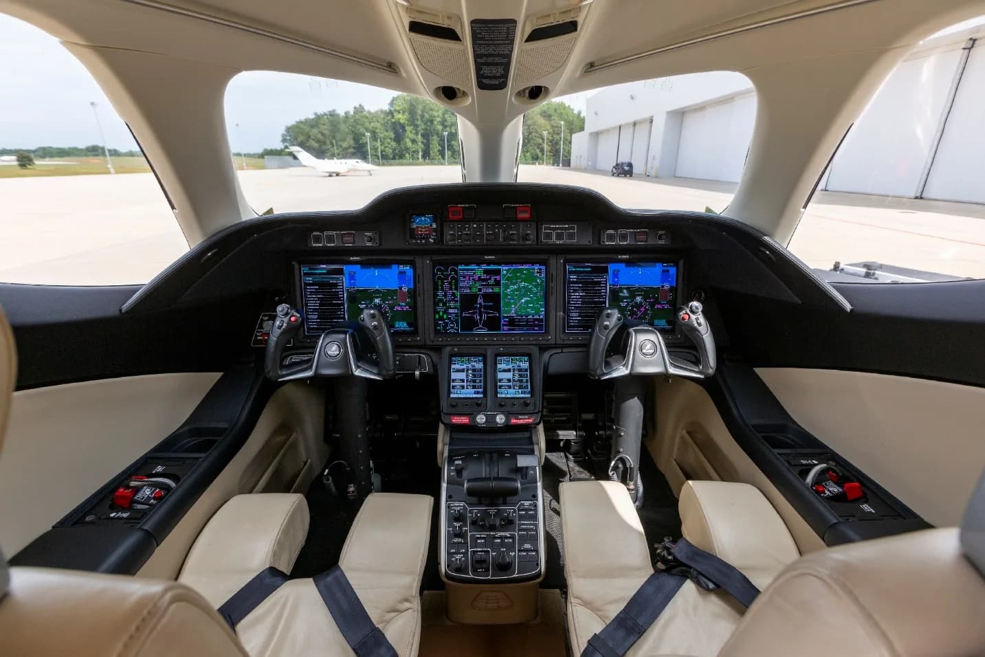 A view from the pilot's seat showing the modern glass cockpit with multiple displays and controls, with an airfield visible through the windshield.