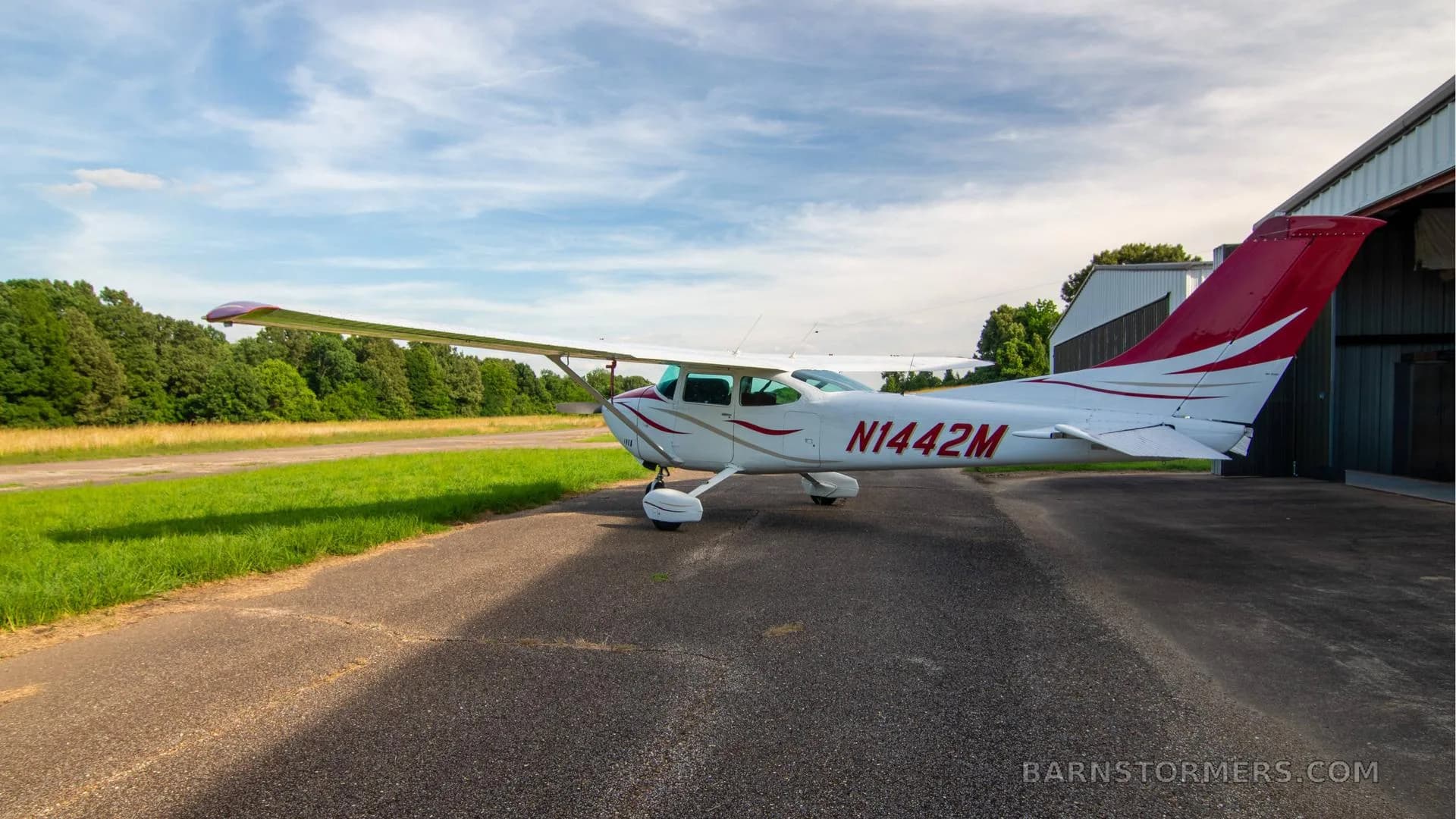 A white single-engine Cessna aircraft with red and gold stripes is parked on an asphalt ramp next to a hangar.
