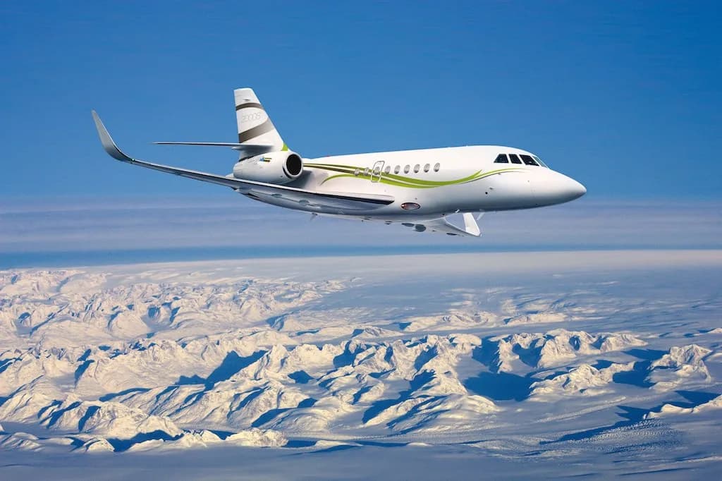 A white Dassault Falcon 2000S business jet with green and grey livery flies over snow-covered mountains under a clear blue sky.