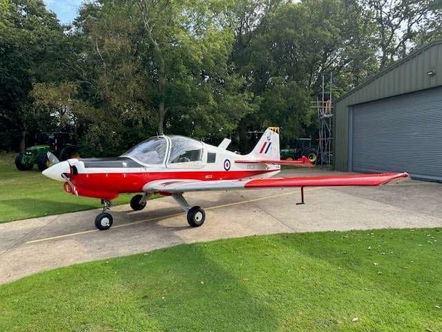 A red and white Bulldog aircraft parked on a paved area near a hangar and trees.