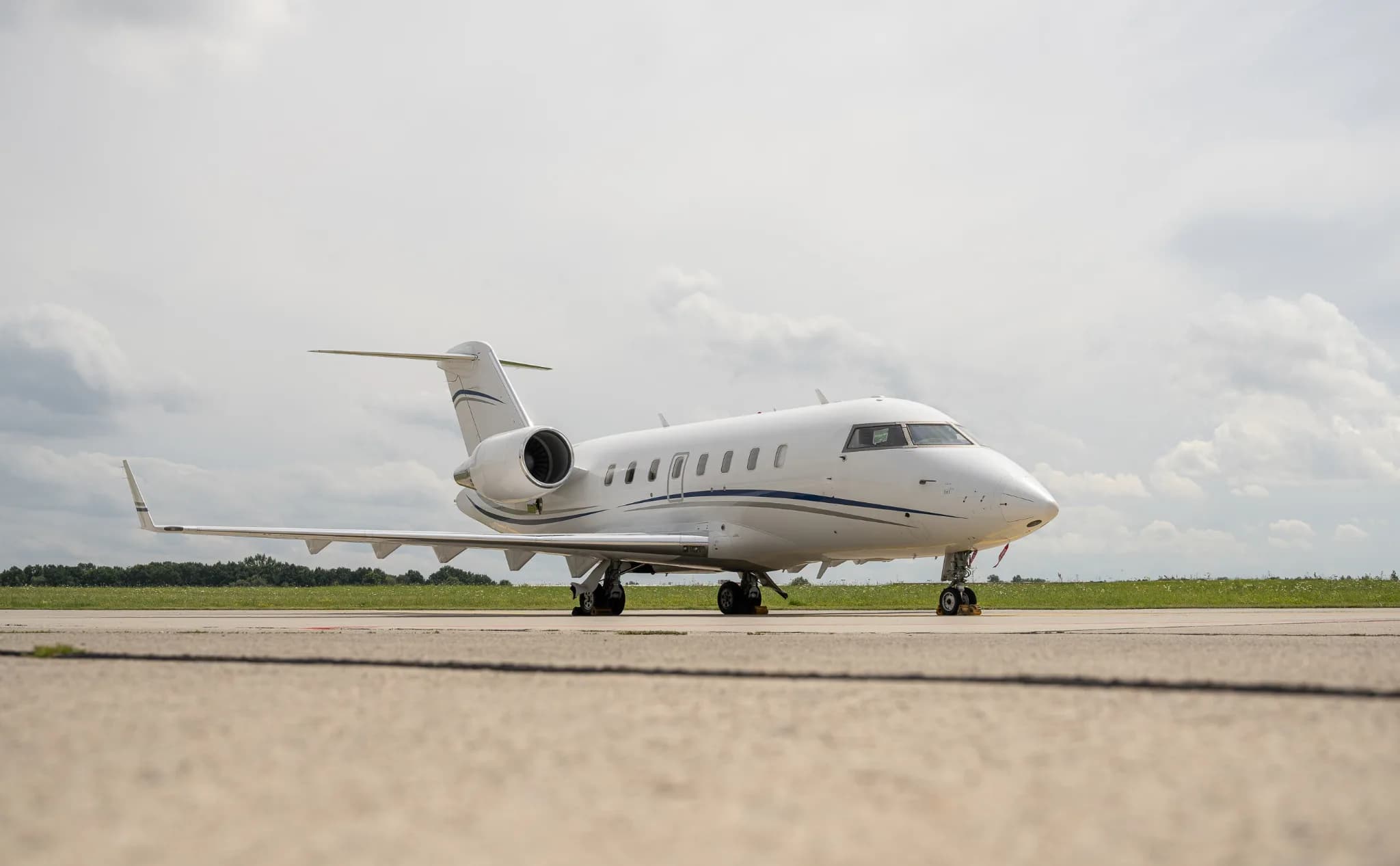A white Bombardier Challenger business jet with blue and grey stripes on the tarmac under a cloudy sky.