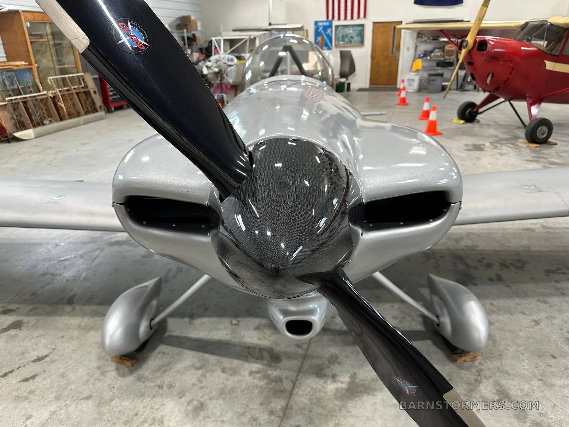 The front view of a silver single-engine aircraft with a black propeller, parked inside a hangar.