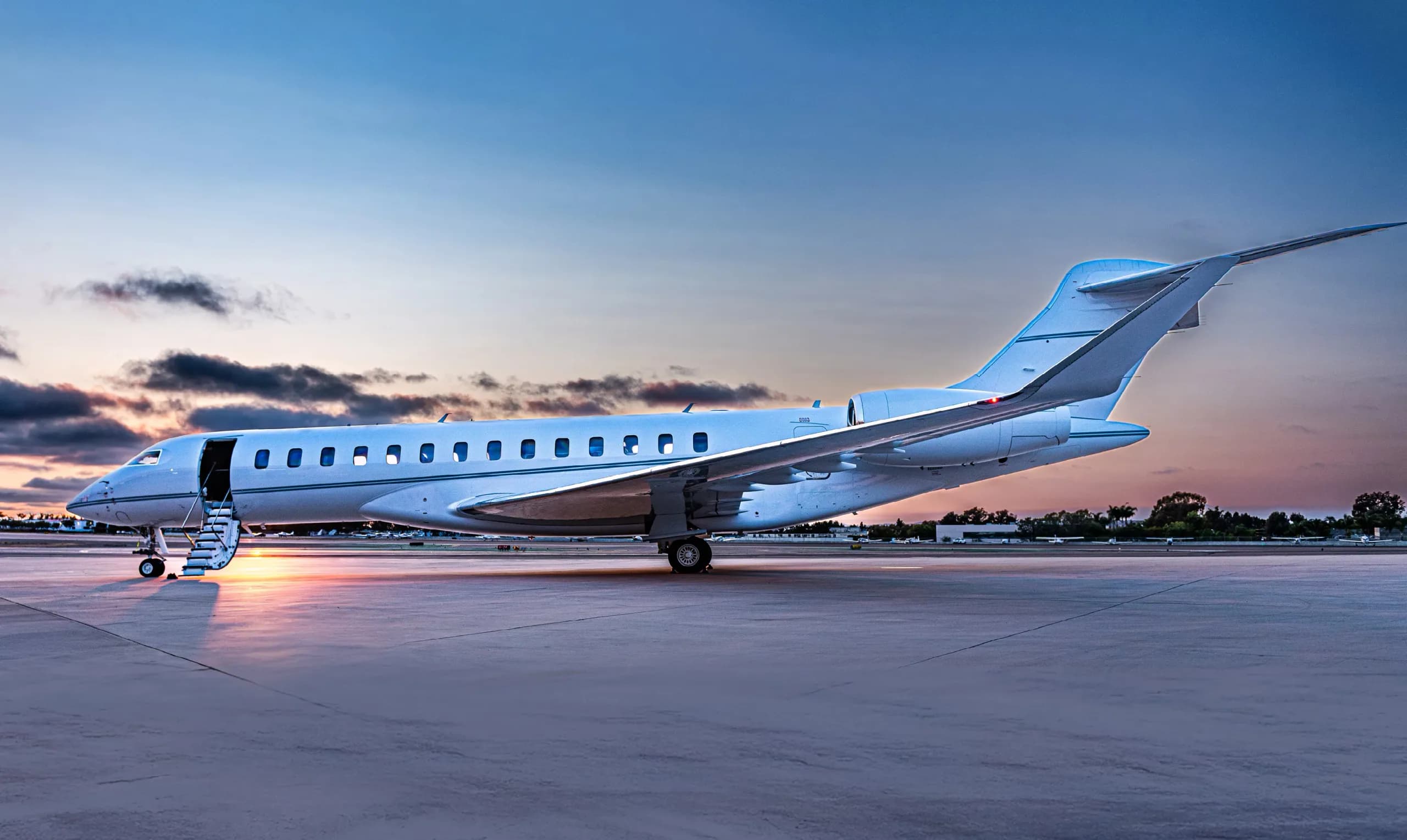 A white Bombardier Global 6000 private jet with its door open and stairs extended, parked on a tarmac at sunset.