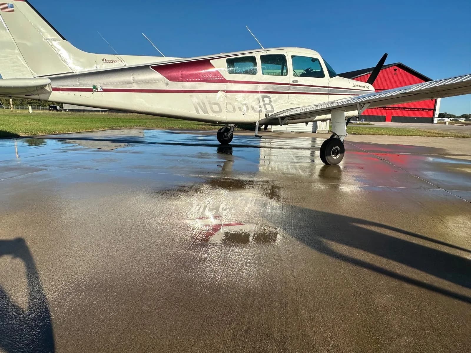 Side view of a white and maroon Beechcraft Sundowner 180 on a wet tarmac, reflecting the aircraft and sky.