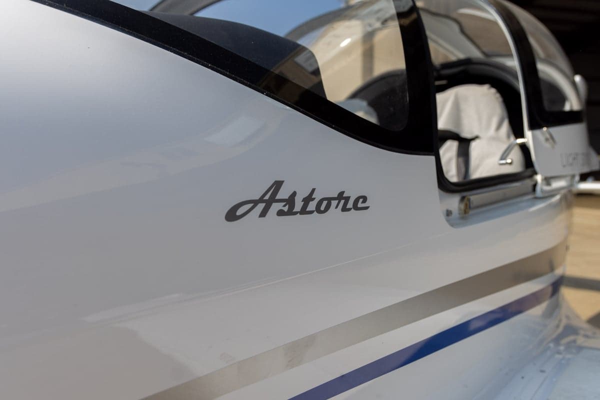 A close-up of the white fuselage of a Tecnam Astore aircraft, showing the "Astore" logo and blue and grey stripes.