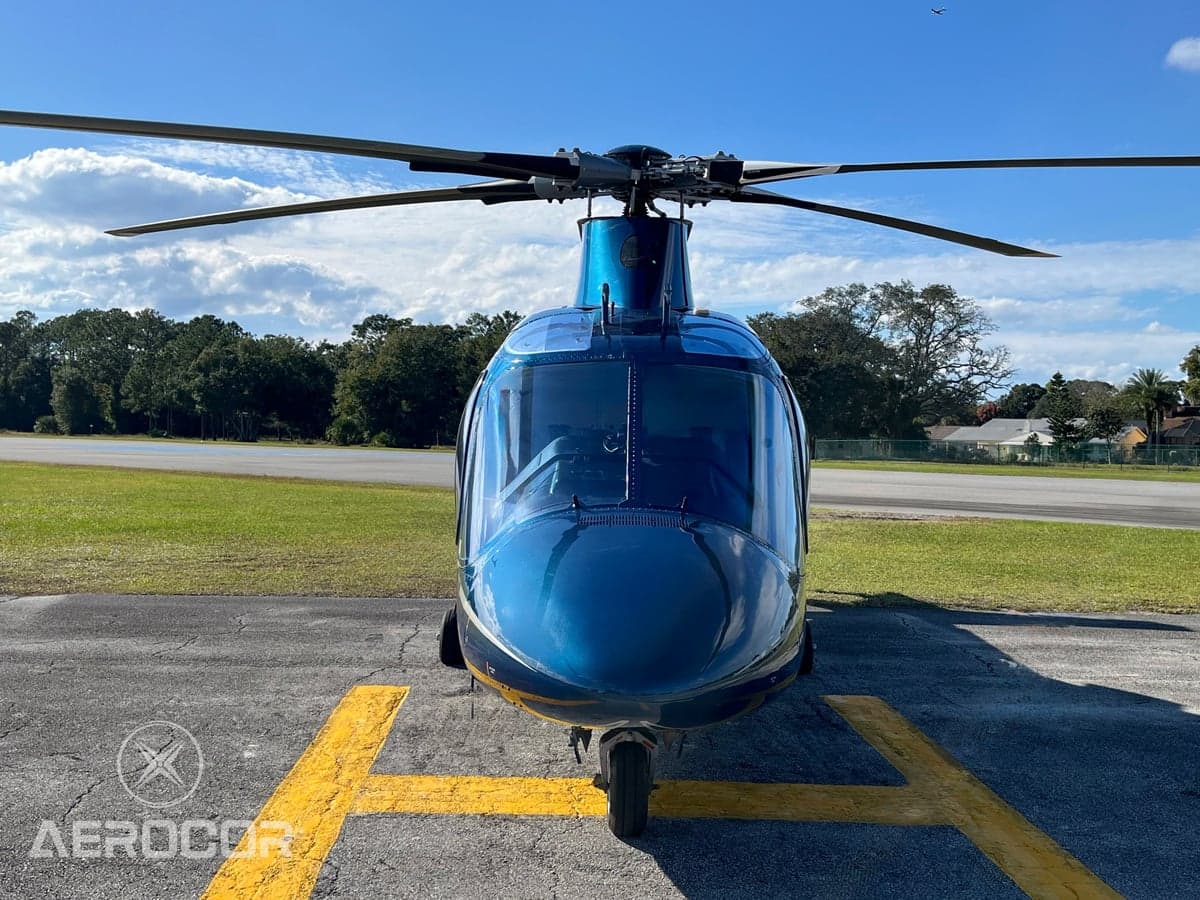 Front view of a blue AgustaWestland AW109 helicopter parked on an airfield under a blue sky.