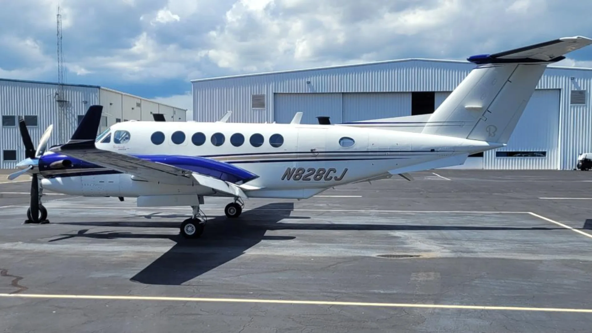 A white and blue Beechcraft King Air N828CJ parked on an asphalt tarmac next to hangars under a cloudy sky.