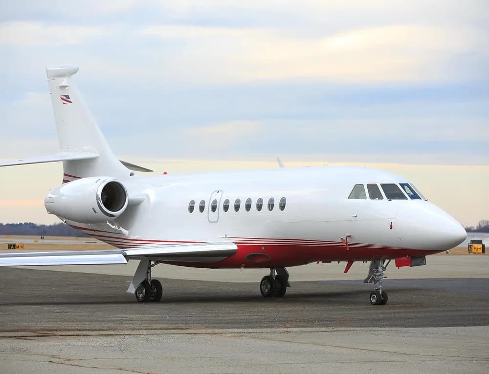 A white and red Dassault Falcon 900 private jet parked on an airport tarmac under a cloudy sky.