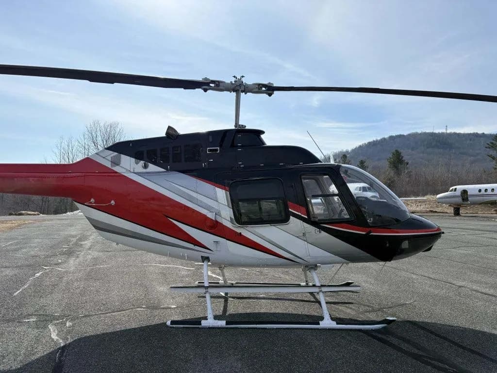 A side view of a red, black, and white helicopter with skids on an airfield.