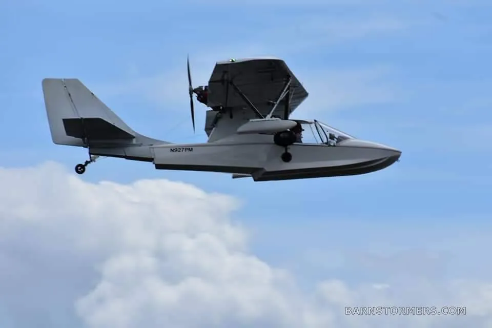 A white experimental seaplane with registration N927PM is flying against a blue sky with clouds.