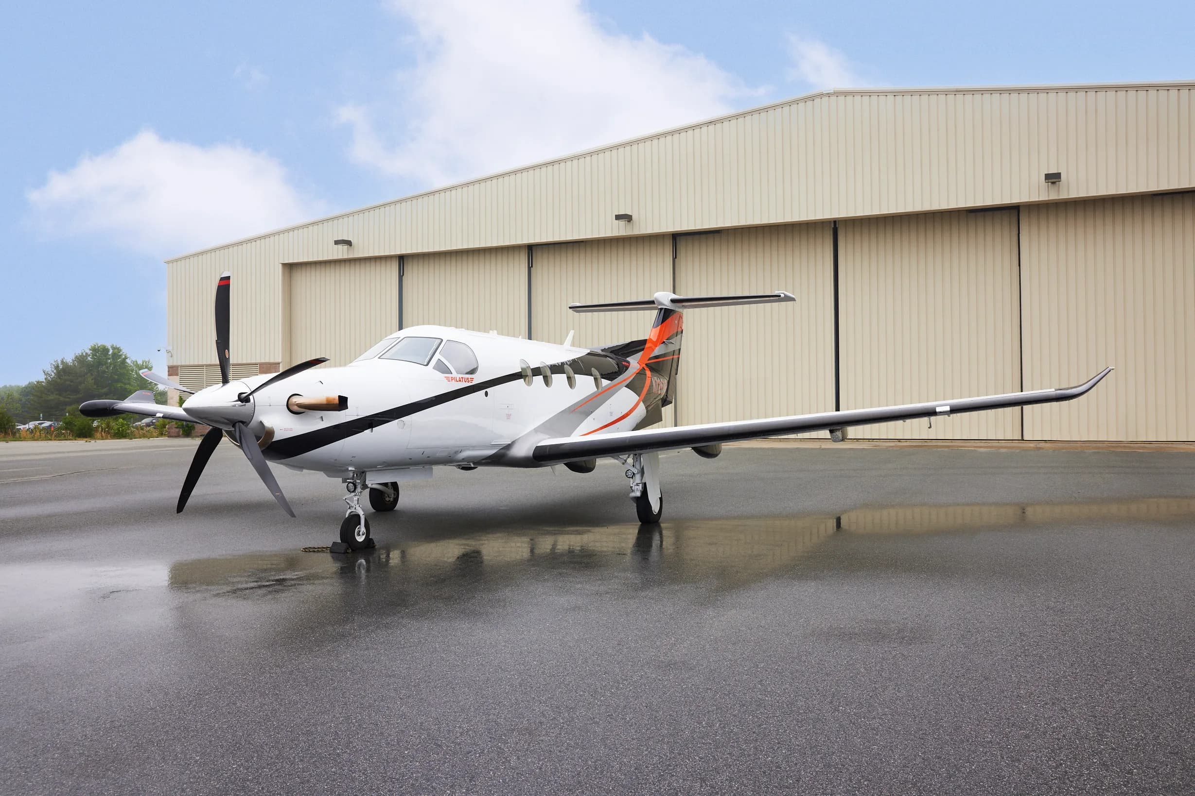 A white Pilatus PC-12 aircraft with black and orange accents parked on a wet tarmac in front of a hangar.