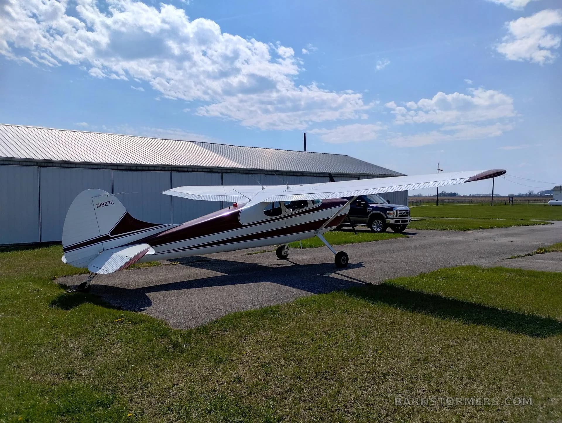 A red and white single-engine propeller aircraft with tail number N1827C is parked on an asphalt pad with a truck and hangar in the background.