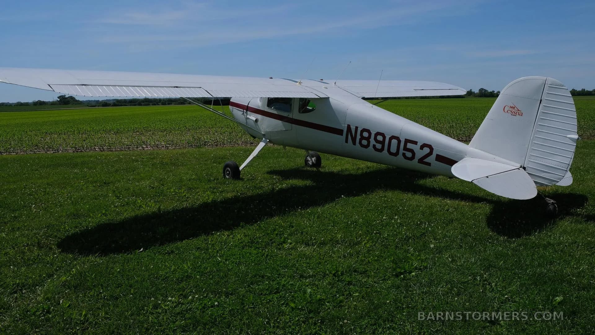 A rear-quarter view of a white Cessna 170 with a red stripe, registration N89052, parked on green grass next to a field.