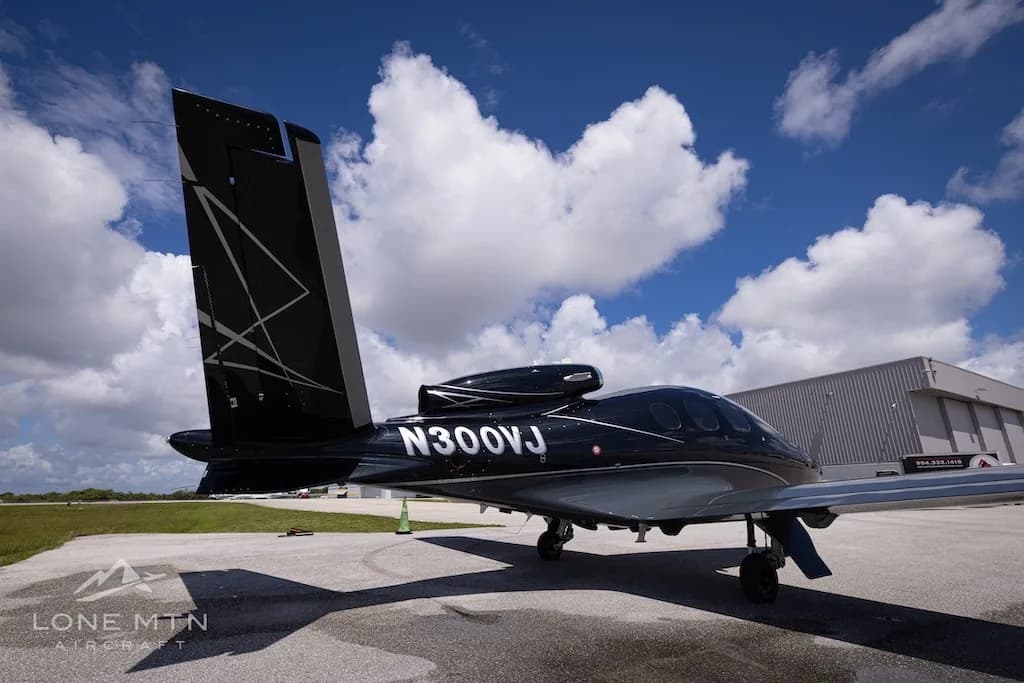 A black Cirrus Vision Jet (N300VJ) with silver accents parked on an airport tarmac next to a hangar, viewed from the rear-left.