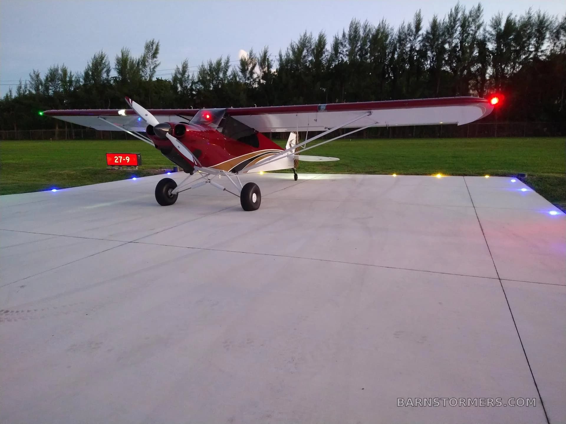 A red and white CubCrafters aircraft with navigation lights on, parked on a concrete pad with ground lights at dusk.