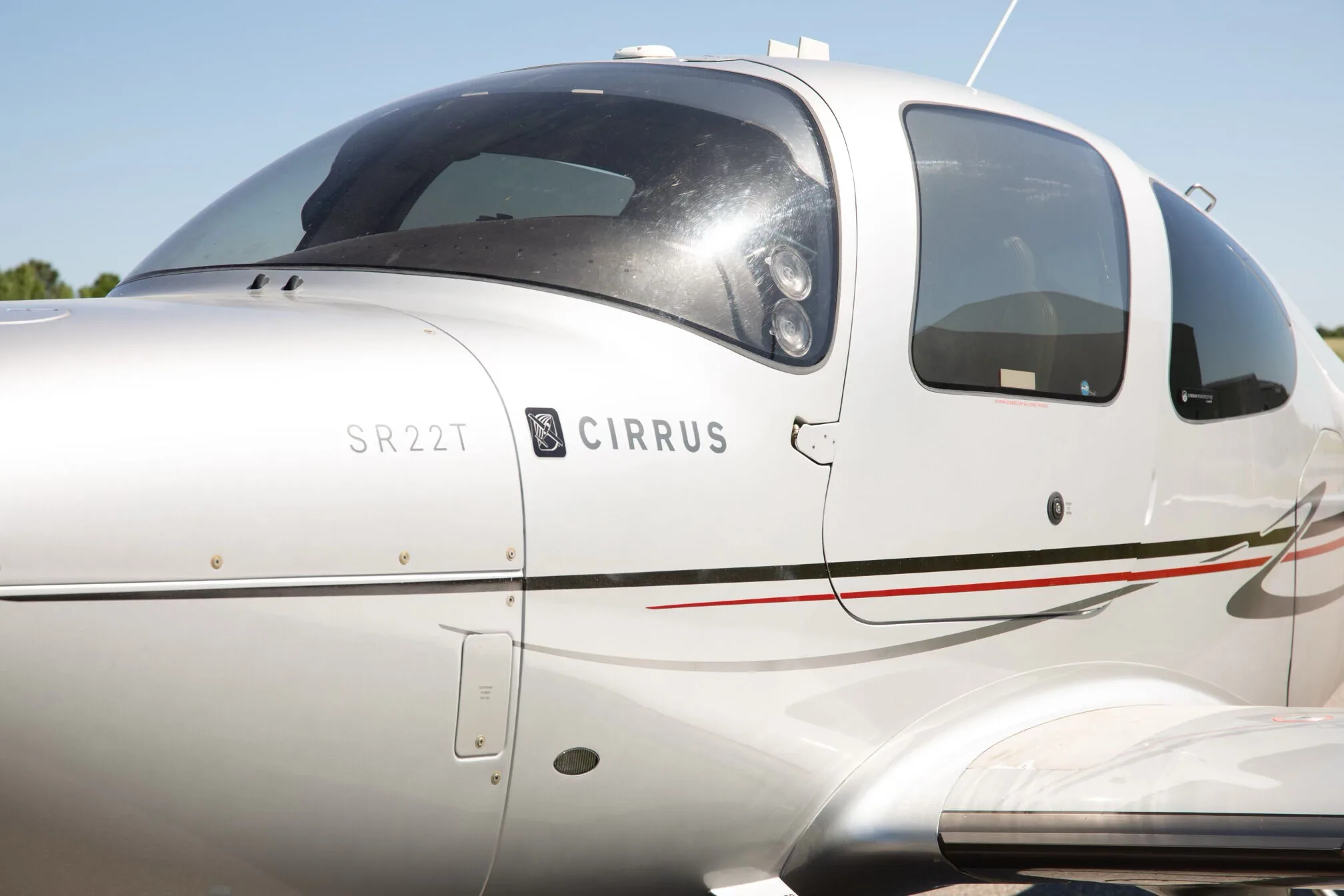 A close-up view of the cockpit windows and side fuselage of the silver Cirrus SR22T aircraft, showing the 'Cirrus SR22T' branding.