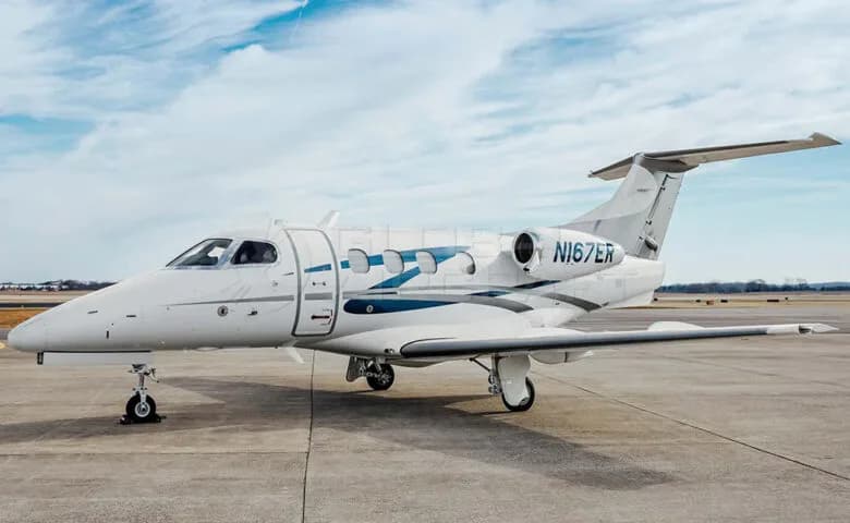 A white Embraer Phenom 300 business jet with blue and grey stripes on the fuselage, parked on an airfield under a partly cloudy sky.