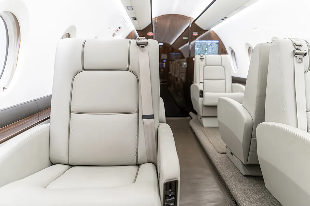 A close-up view of a light-colored leather aircraft seat with a visible seatbelt, looking down the cabin aisle.
