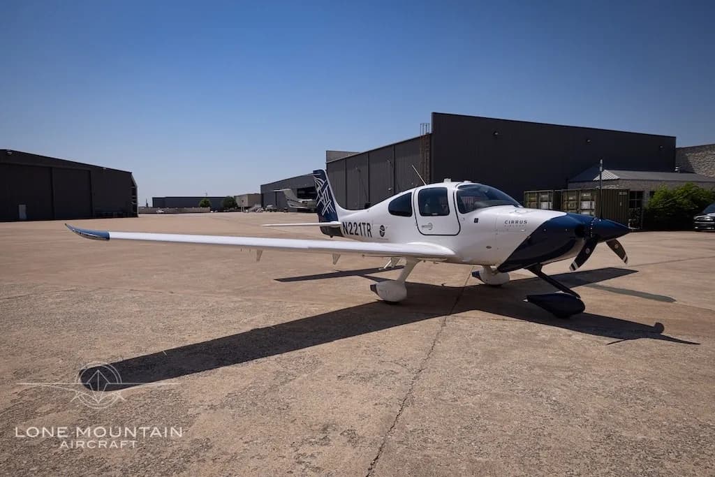 A white Cirrus SR22 aircraft with a dark blue nose and tail, registration N221TR, parked on an airport tarmac.