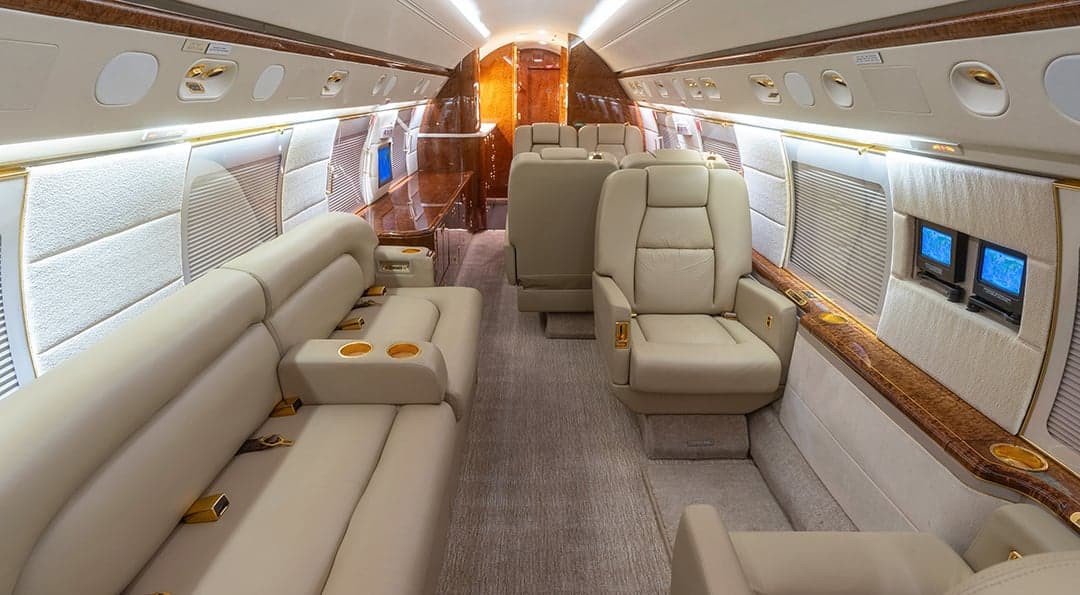 A private jet cabin with a beige leather couch, individual seats, and polished wood paneling, looking towards the rear.