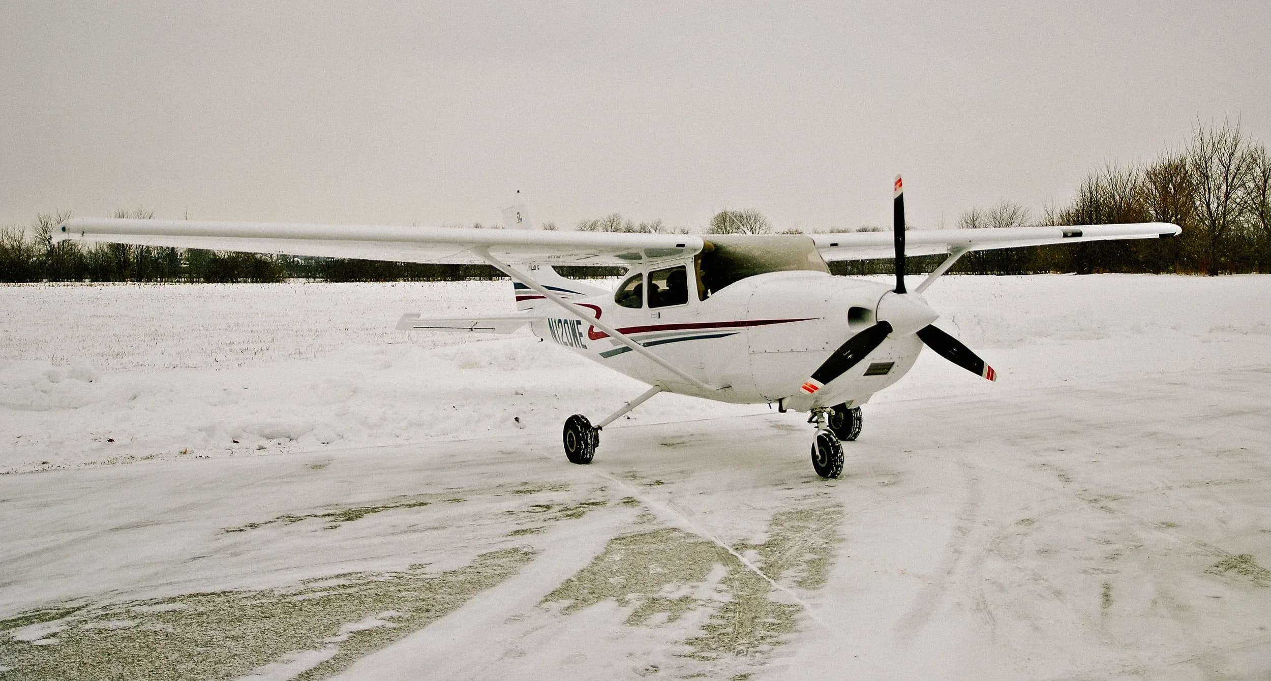 A white single-engine high-wing aircraft with red and blue stripes is parked on a snow-covered field under an overcast sky.