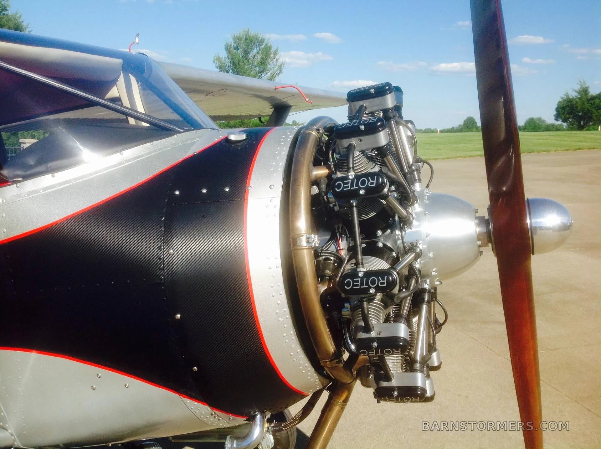 A close-up view of the exposed Rotec radial engine and wooden propeller of a silver and black aircraft.