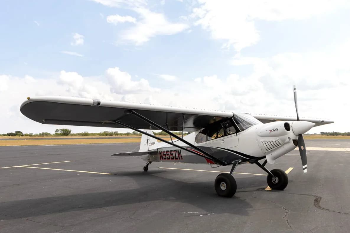 A three-quarter front view of the white and grey X-Cub aircraft N555ZM with red accents, parked on an asphalt runway.