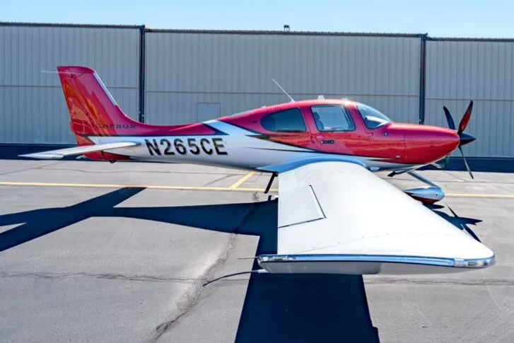 A side view of the red and silver Cirrus SR22T GTS Carbon aircraft, registration N265CE, parked in front of a hangar.