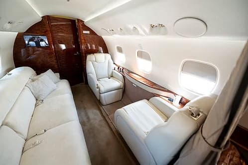 The rear section of a private jet cabin featuring a long cream sofa, individual seats, and polished wood accents.