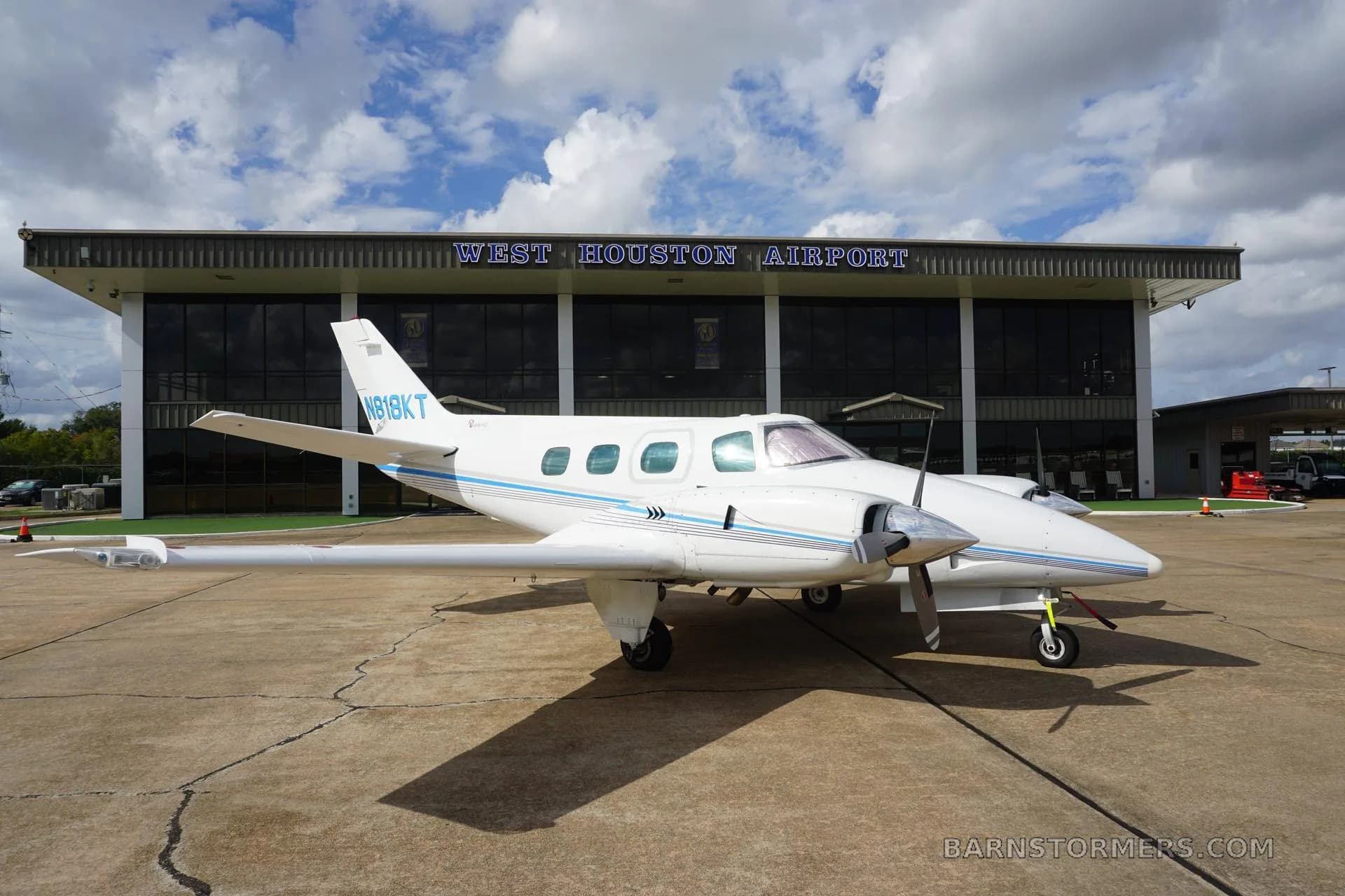 A white twin-engine propeller aircraft with blue stripes (N818KT) is parked on the tarmac in front of the West Houston Airport building under a cloudy sky.