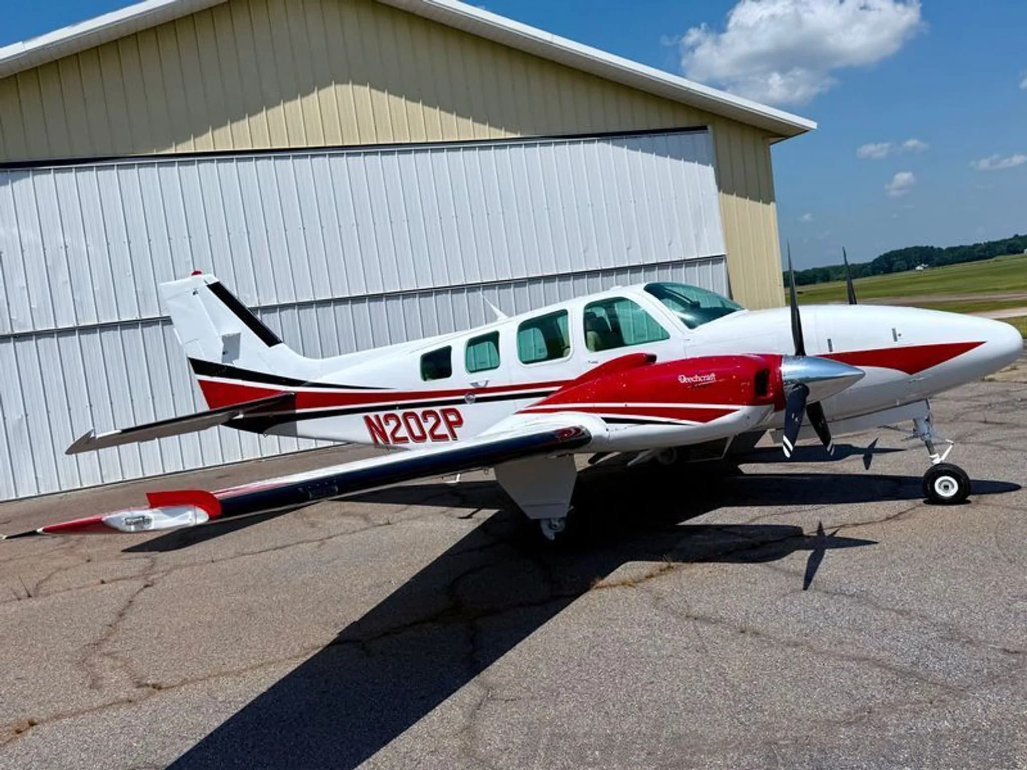 A white Beechcraft Baron with red and black stripes, N202P, parked on an asphalt ramp in front of a hangar.