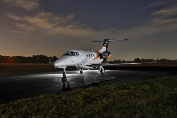 A white private jet with orange and black accents sits on a wet tarmac at night, illuminated by its landing lights.