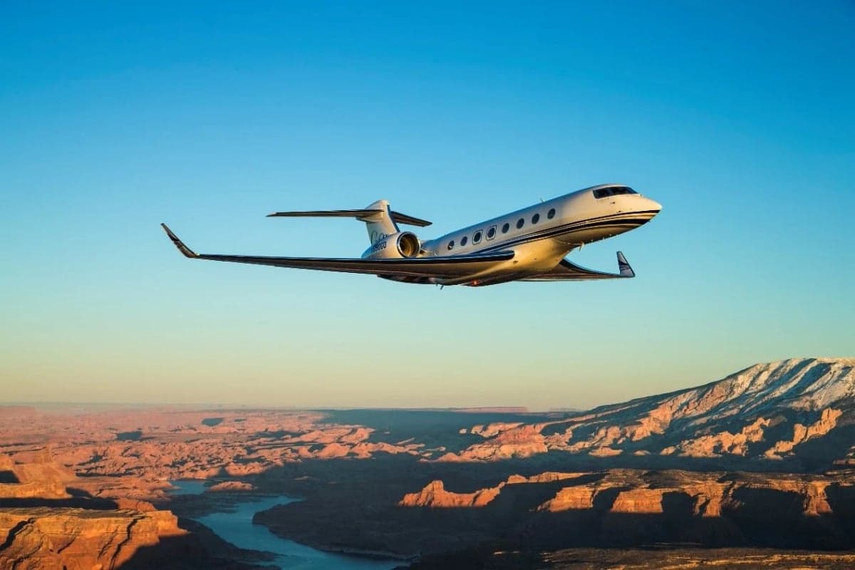 A white Gulfstream G650 business jet flying over a vast canyon landscape at sunset.