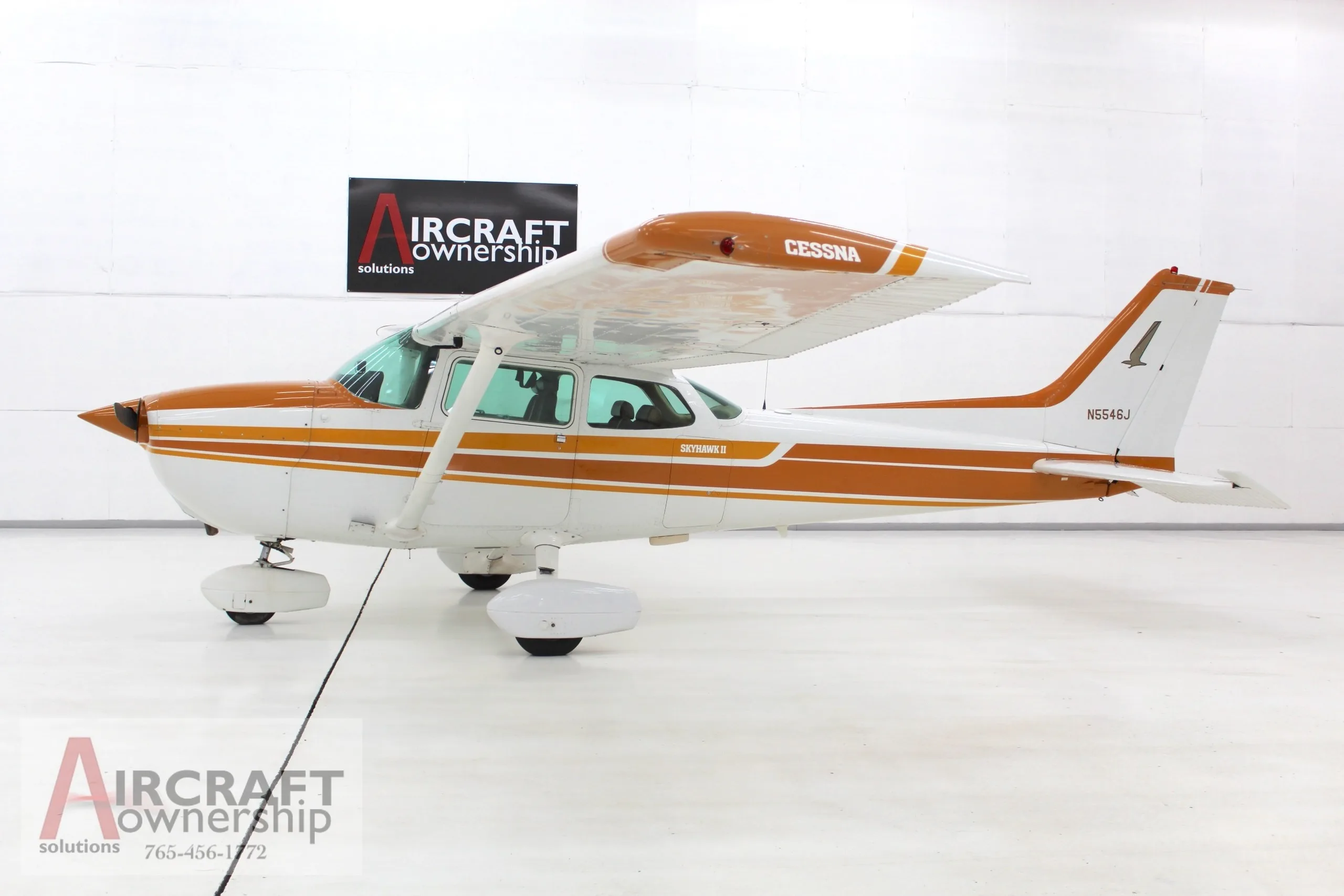 A white and orange Cessna Skyhawk II (N5546J) is parked in a brightly lit hangar, viewed from the side.