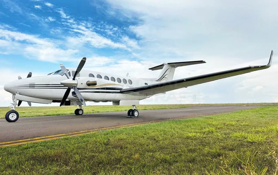 A white Beechcraft King Air twin-turboprop aircraft with black and gold stripes is parked on a runway under a partly cloudy sky.