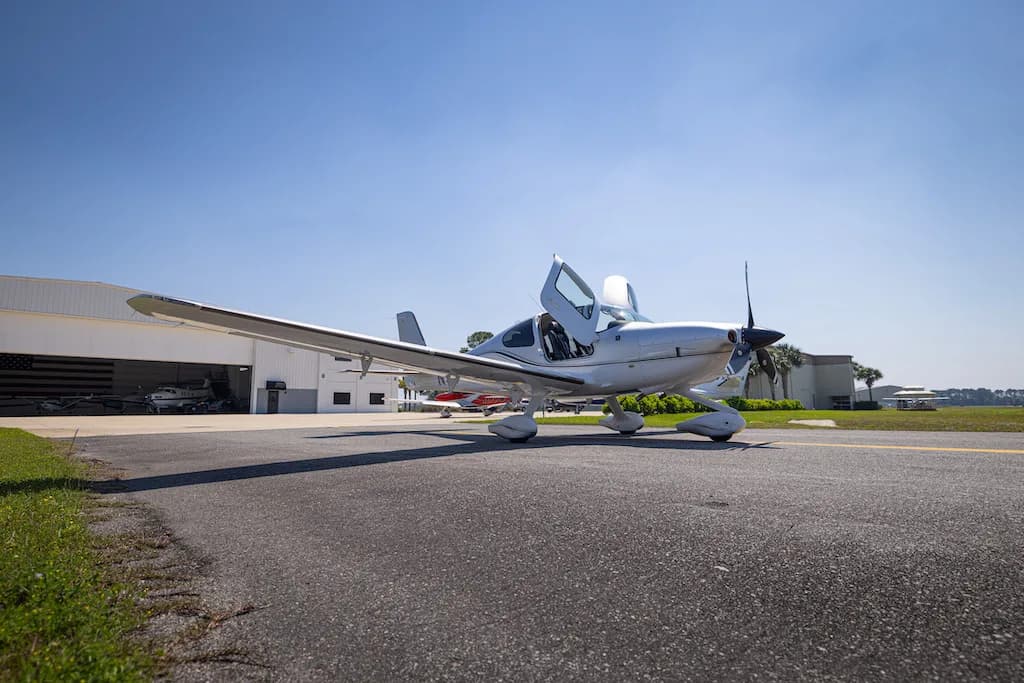 A silver Cirrus SR22 aircraft with its doors open, parked on an airfield tarmac with hangars in the background.