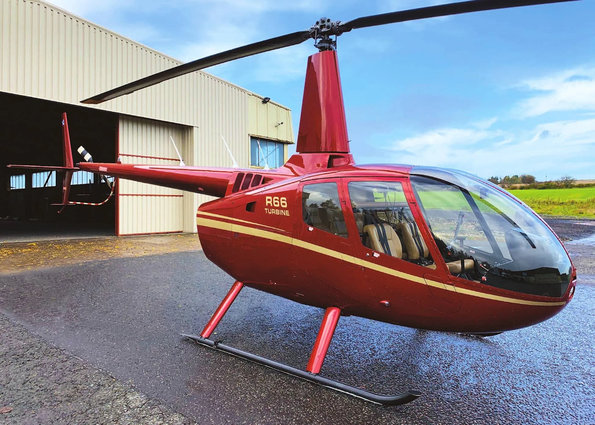 A red Robinson R66 Turbine helicopter with gold stripes parked on a wet tarmac next to a hangar.