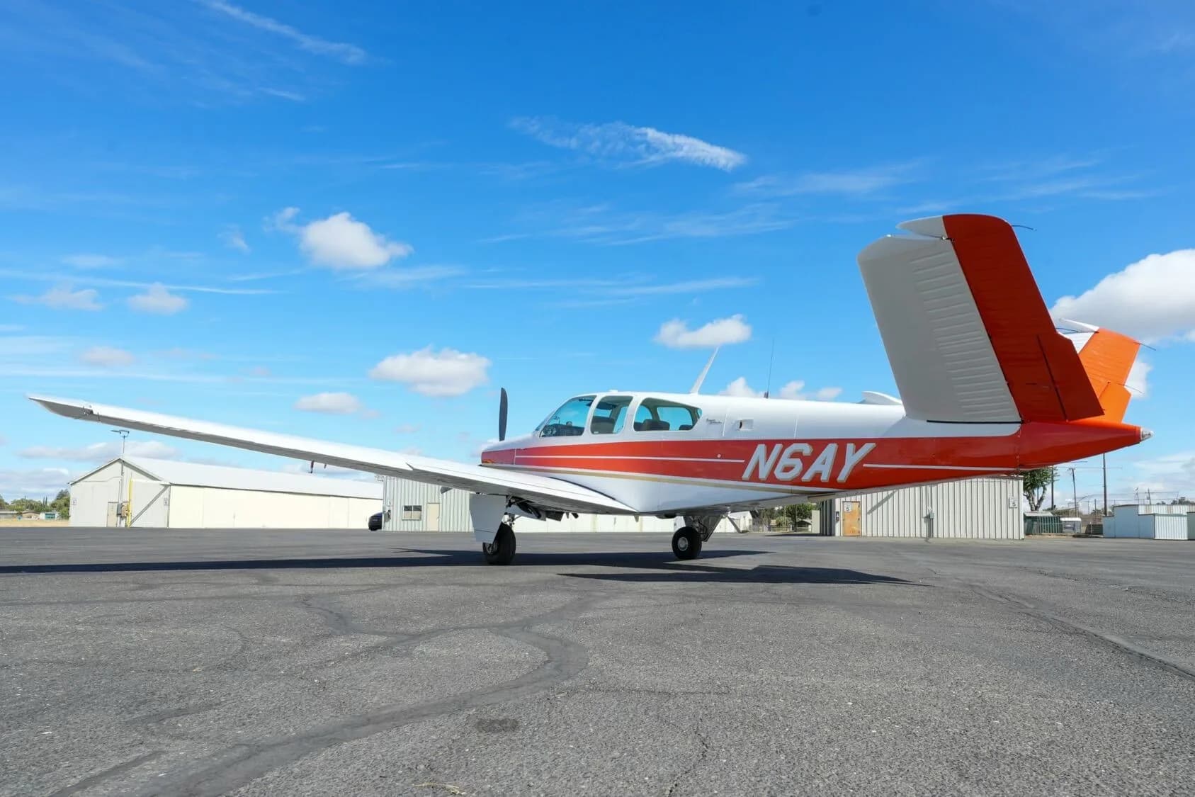 A side view of the white and orange Beechcraft Bonanza N6AY parked on an airport tarmac.