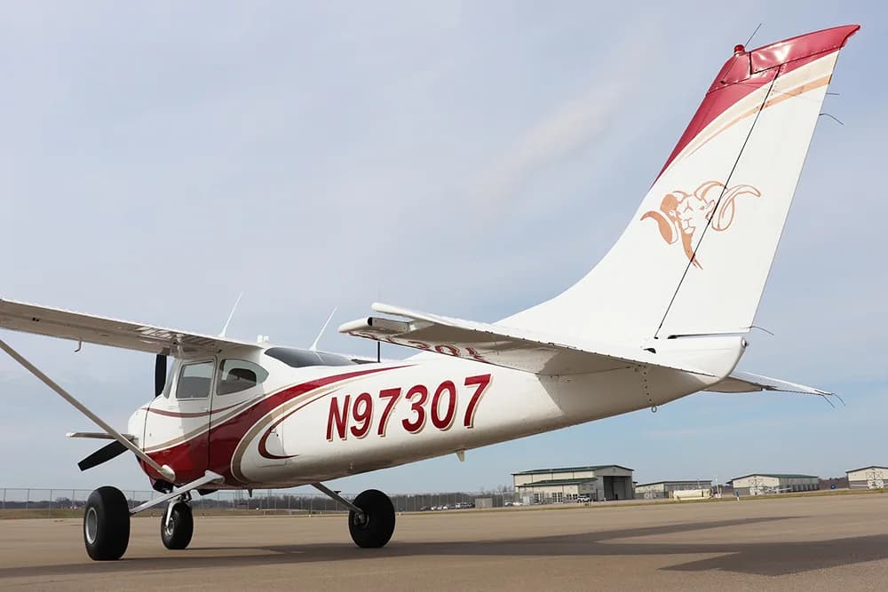 A rear-side view of a white and red single-engine high-wing aircraft, N97307, parked on an airfield with hangars in the background.